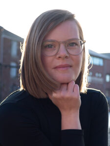 person with glasses and bobbed hair with head resting on hand, buildings in background