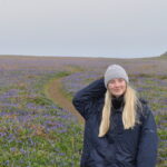 Alice with long blonde hair, a grey cap and dark jacket. Standing in a misty landscape. 