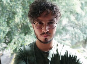 person with glasses, curly hair and facial hair in front of trees