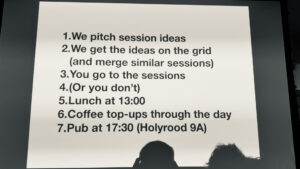 Photo of a slide showing the high level plan for the day: 1 pitching sessions, 2 get ideas into a grid and merge similar, 3 go to sessions, 4 or you don't, 5 lunch at 1pm, 6 coffee throughout the day, 7 pub at 5:30