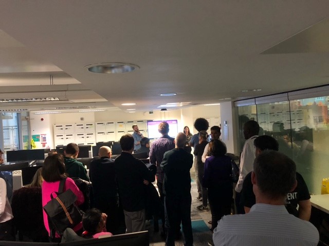 A Large room of people watch a presentation while stood around desks