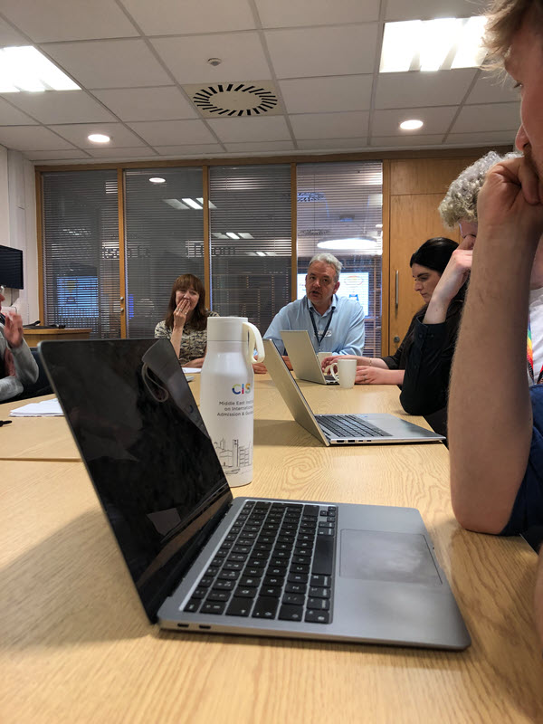 A group of staff sit at a table in front of laptops while discussing.
