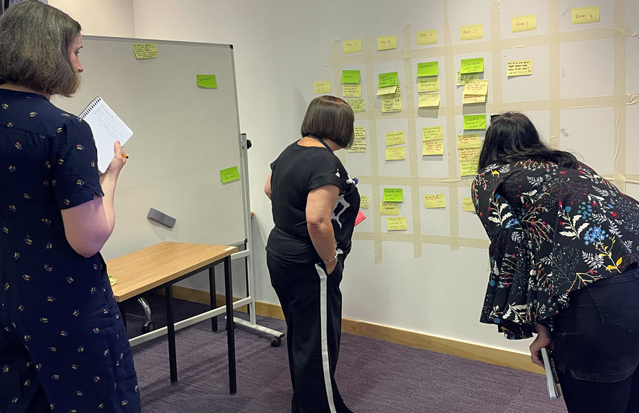 Three people look at a timetable contructed with post-it notes on a wall.