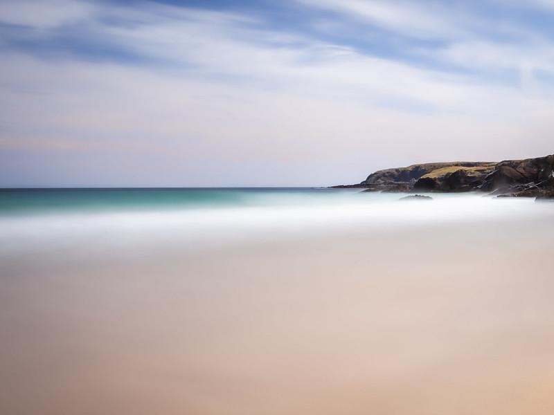 An image of a white sandy beach with turquoise water and rocky cliffs in the background. The image is taken using a long shutter speed, making the waves blur into the sand. The image is of Port Ness beach on the Isle of Lewis in Scotland. 