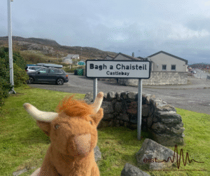 Image Description: A stuffed toy Highland Cow facing a road sign. The sign reads ‘Bàgh a Chaisteil, Castlebay,’ with a stone wall and a grassy area nearby. In the background, there is a modern car, some buildings, and rocky hills under a cloudy sky. 