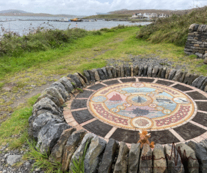 Image Description: A circular stone mosaic set into the ground and surrounded by upright stone slabs. The mosaic is detailed with colorful, intricate patterns and symbols. It is designed to be a compass that features motifs of the island. On the side closest to the camera sits a stuffed animal toy. Around it is a grassy area with some rocks, leading towards a body of water in the background.