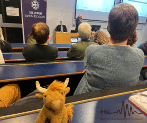 Image Description: Photograph of a lecture hall. Several people are sitting in tiered seats facing the front, where a person is giving a presentation at the podium. There is a blue banner at the front with text and a logo, reading ‘University of Edinburgh’ in both English and Gaelic. A plush toy cow with light brown fur and white horns is visible on the desk in the foreground facing the camera.