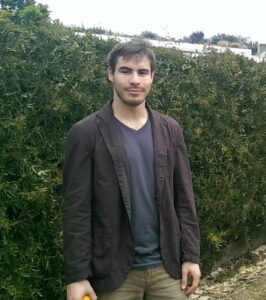 Drew standing in front of a hedges smiling at the camera. He wears a black jacket with a blue shirt underneath