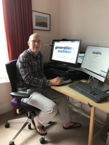 David Porteous sat at his desk with monitors displaying Generation Scotland and CovidLife logos