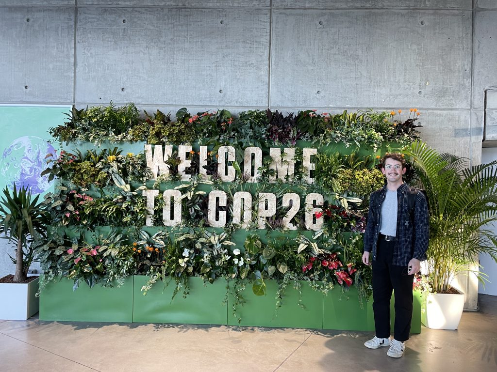 The author of the blog post is standing next to the welcome sign at the COP26 'Green Zone'. The sign is a living art exhibit constructed out of a variety of plants and it reads "Welcome to COP26".