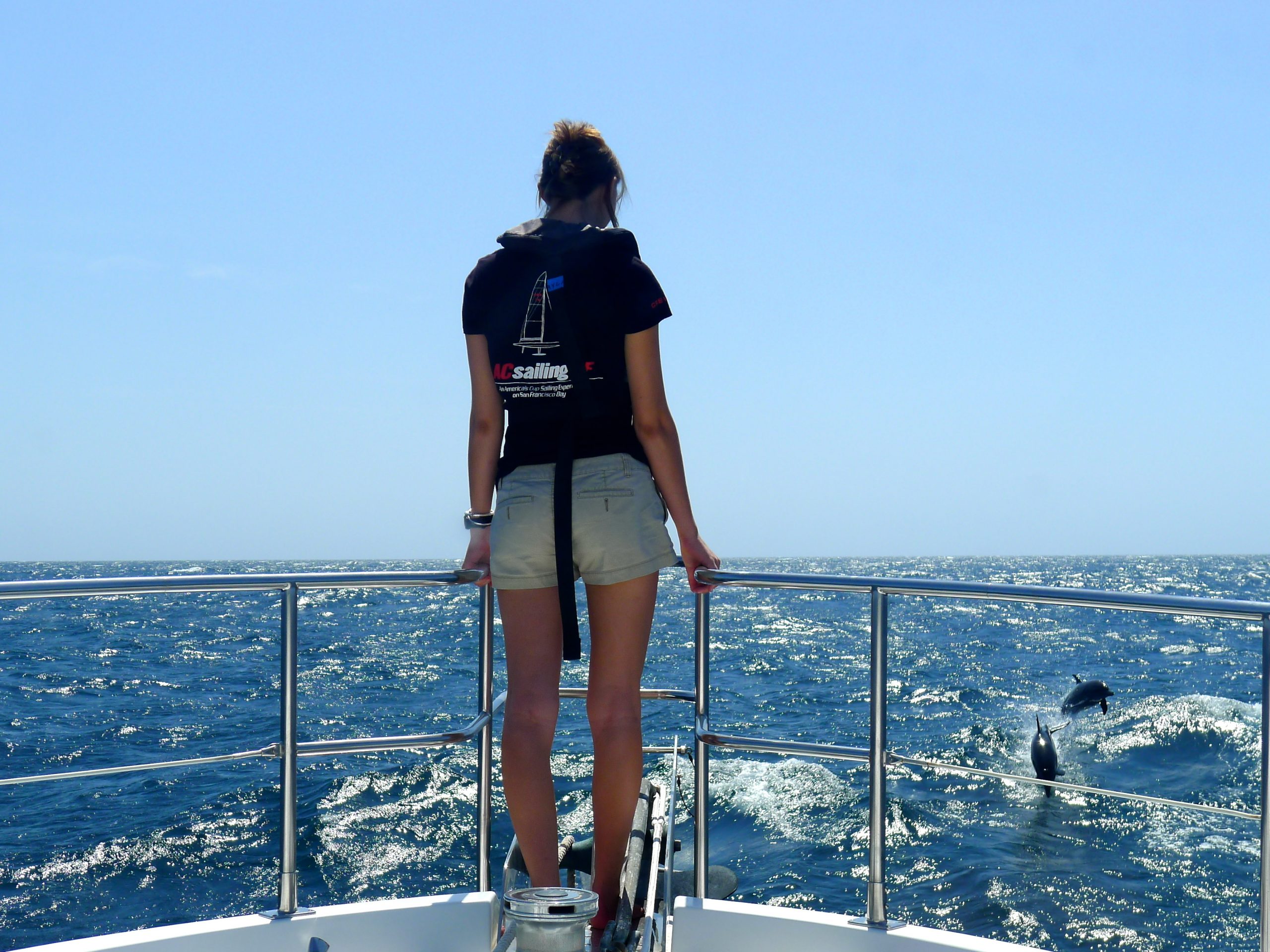 Ayla stands on a boat's deck with her back to the camera, looking out towards the ocean. The sun shines brightly in the sky, casting a warm glow on the woman and the surroundings. In the background, the ocean sparkles and two dolphins can be seen leaping in and out of the water. The woman is wearing shorts and appears relaxed and content as she enjoys the beautiful scene.