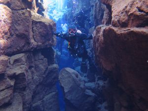 Ayla is scuba diving in Iceland between the tectonic plates of Eurasia and North American. The water is a deep blue color and she is wearing a full wet suit, goggles, flippers and an oxygen tank. Ayla can be seen touching both plates with her arms outstretched as she swims between the towering rock walls.