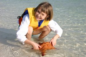 Ayla, at age 7, is crouching in shallow water wearing a yellow life vest in the Bahamas. She is holding an orange sea star in her hand that she rescued from above the tidal line. The sea star appears to be healthy and intact.