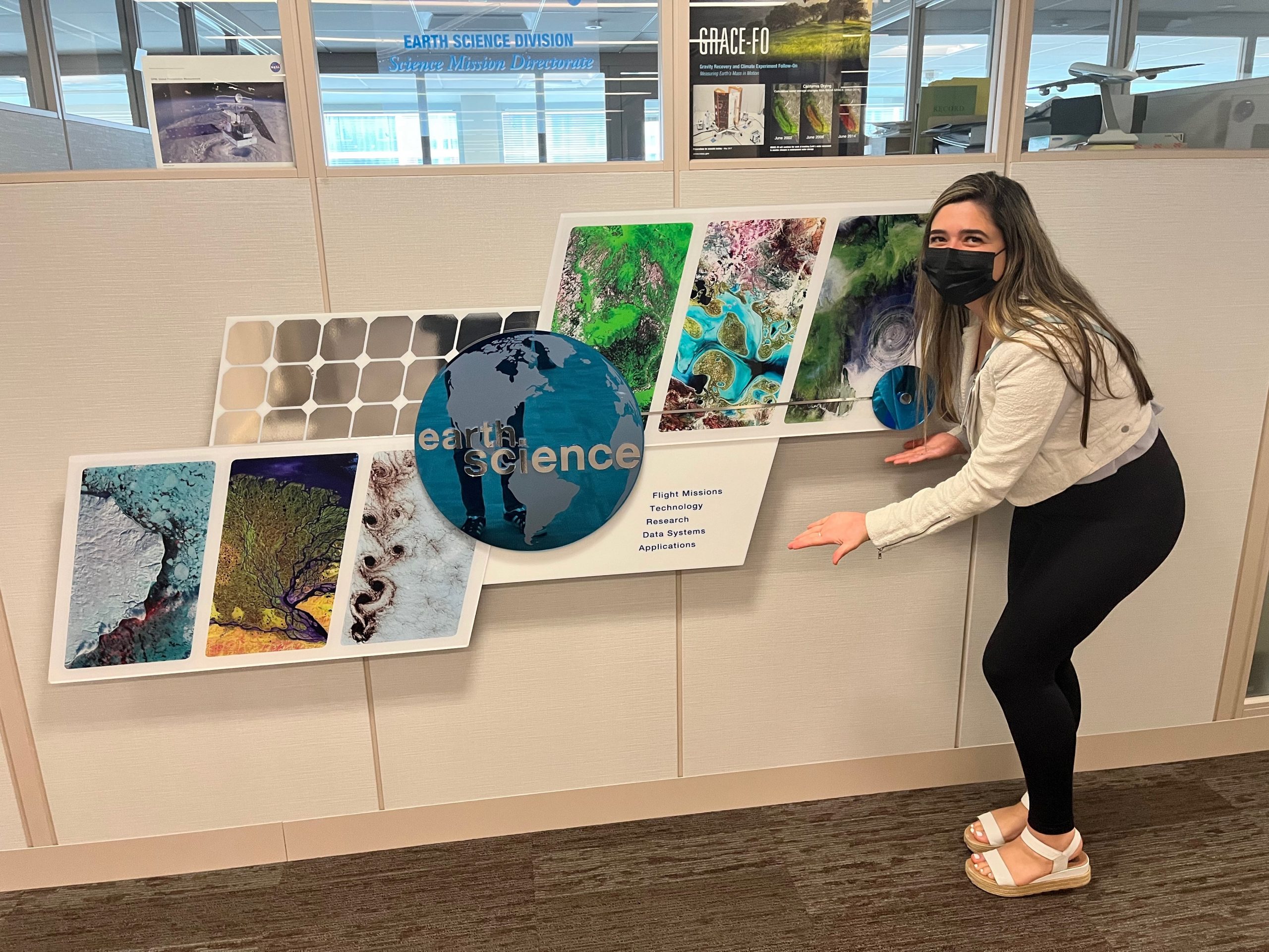 Mia, a NASA intern, stands in the office at NASA HQ, pointing at the Earth Science Division sign. She's dressed casually in a denim jacket, leggings and sandals.
