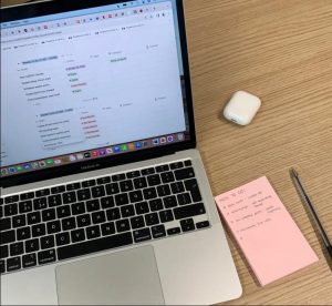 A photo of Emmy's desk. Her Macbook screen shows her to-do list on Notion. There's a stack of pink sticky notes next to it with more to-do list points.