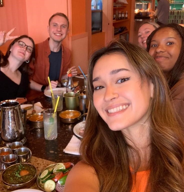 A selfie of Emmy with the rest of the communications and marketing team in the background. Everyone is sitting around a table in a restaurant.