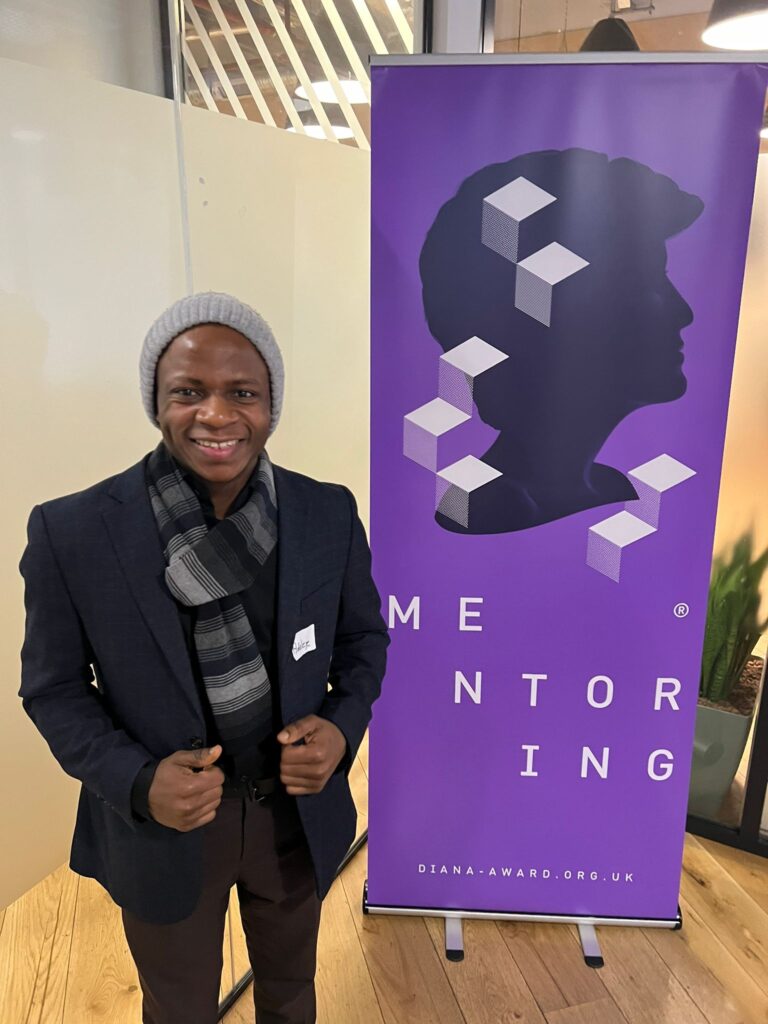 Photograph of Ademiku standing in front of the Diana Mentoring Award banner.