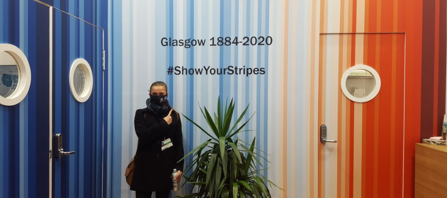 Alumnus Amy stands in front of a Climate Stripes background that states "Glasgow 1884 - 2020 #ShowYourStripes"