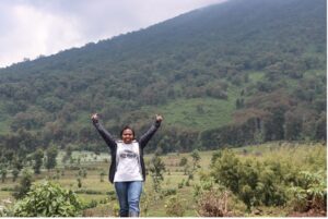 Jeannette poses for a photo with arms raised, in front of a mountain with lush greenery.