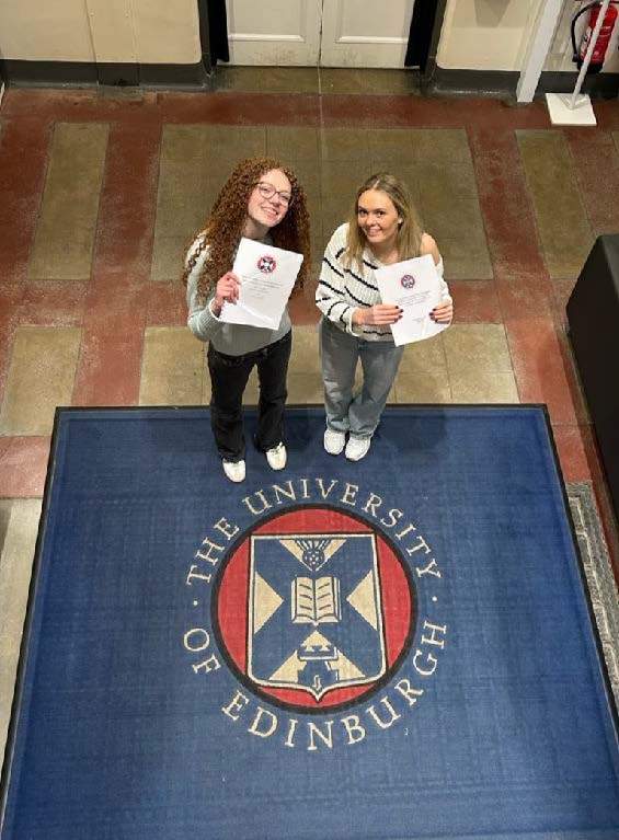 Ellis poses with a friend holding their dissertations, and standing next to a carpet with the University of Edinburgh logo.