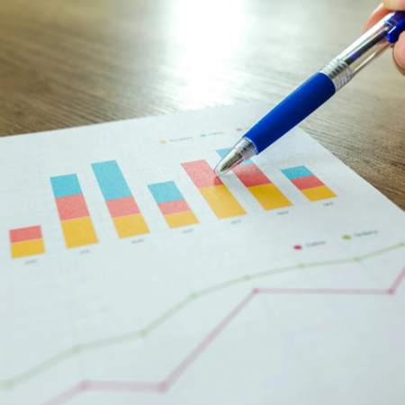 Stock photo showing a printout of a colourful stacked bar chart above a line graph. The printout is placed on a wooden desk, and a person’s hand is pointing a pen at one of the bars.