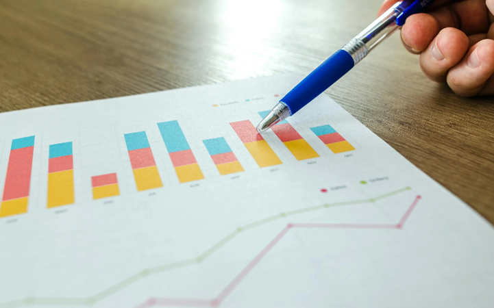 Stock photo showing a printout of a colourful stacked bar chart above a line graph. The printout is placed on a wooden desk, and a person’s hand is pointing a pen at one of the bars.