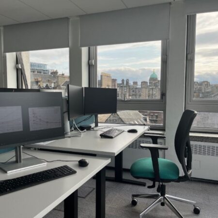 In the foreground, the workspaces on floor K of Argyle House with a monitor displaying multiple graphs. In the background, a view of Edinburgh rooftops through the floor K windows.