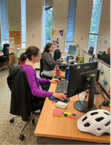Two interns sat at desk in a busy office environment.