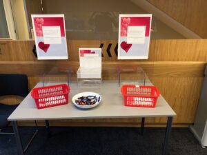 Table with 3 transparent small stands. There are 2 read trays with pieces of paper in them. The is a small bowl with chocolates