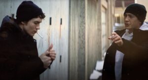 Ilya is smoking while wearing a dark jacket and Shane is in a light grey hoodie, toque, and jacket, gesturing that Ilya shouldn't be smoking there. They are in an alleyway behind a hockey arena.