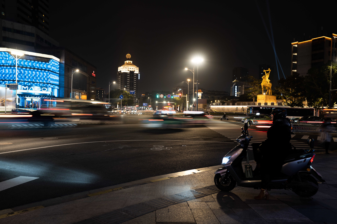 An ebike driver sits parked on an ebike at an intersection at night as blurred traffic flows by. Lights from tall buildings color the background.