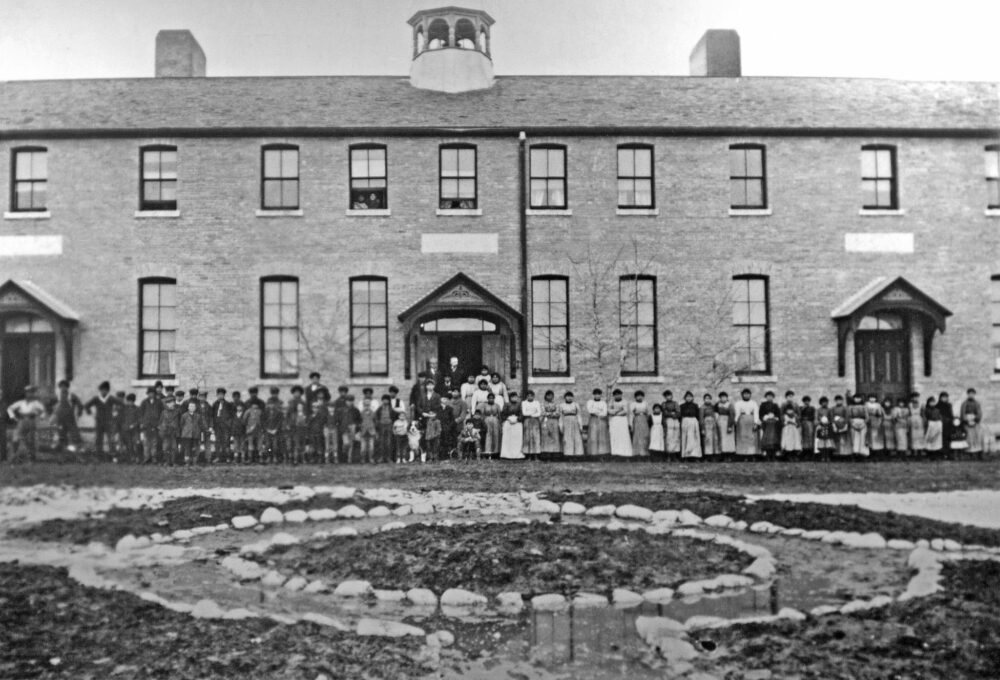 A black and white picture of a large two story stone building with many young students standing in front.