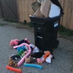 A black garbage bin with a pile of colourful toys in front of it. 