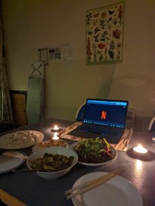 Image shows table set with with plates, chopsticks and candles. Food is in the middle, with a laptop displaying a Netflix logo at the edge of the table.