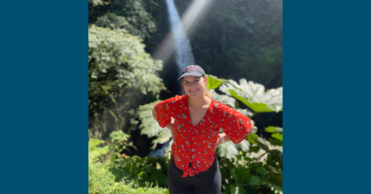 Photo of Megan smiling with a waterfall in the background. Megan is wearing a black cap, and a floral red top with a blue flower motif