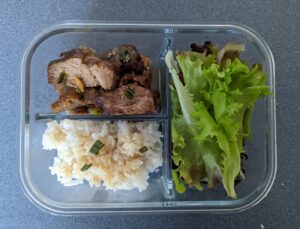 Glass lunchbox with three compartments, each containing rice, salad leaves, and grilled meat