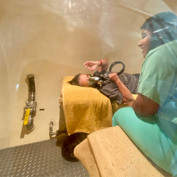 Mallaika in green scrubs speaking to a patient wearing a mask inside the hyperbaric chamber 