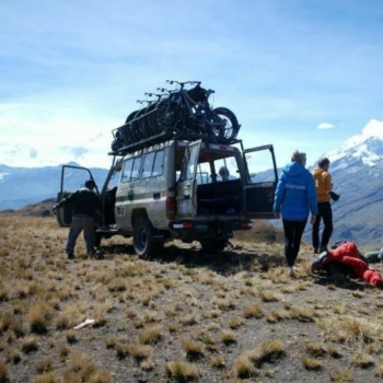 Land rover with bikes on the roof stopped with snowy moutains in background and 3 people standing nearby.