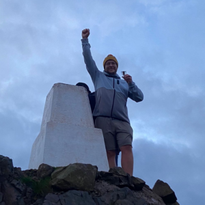 man standing on top of rock with one hand in the air celebrating, other hand holds a little trophy