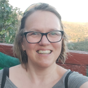 Hilary smiling at camera, wearing glasses and a green top. She has light brown hair. In the background is a fence and trees.