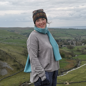 Jess Gurney smiling at camera wearing hat, and scarf with grey jumper. Jess is standing in front of a background of green fields, trees and a river.