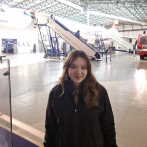 Cassidy wearing a black jacket and long brown hair, inside an air hangar with planes in the background.