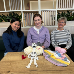 Stefanie, Amberley and Lorna sit at cafe table in front of items they have crocheted. They are in front of a background of shelves and plants.