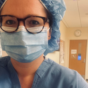 Rachael Forsythe wearing blue scrubs, a face mask and hat. She also has glasses on. In the background is an office door and clock on the wall.