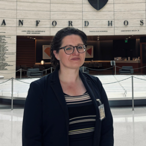 Rachael Forsyth standing in the foyer of Standford Hospital. Rachael has dark blazer on and stripey top, brown hair and glasses. In the background is the foyer wall and a fenced off piece of art.