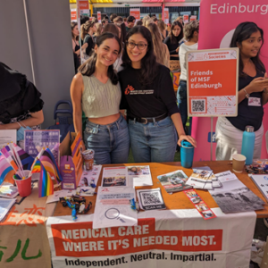 Two students from Friends of MSF stand behind an information tablet with flyers and info on it. They are at a busy student society fair, surrounded by signage and people in background.