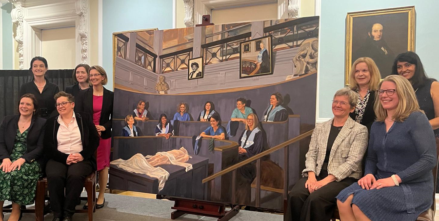 Nine women surgeons (the subjects of the painting) sitting beside the painting 'Eleven Surgeons' by Kirsten Mackinnon. 