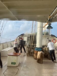View from the deck of a tall ship with three people working near ropes and rigging under cloudy skies.