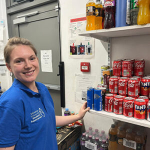 Student working in a shop - smiling at camera with cans of Coca Cola on a shelf