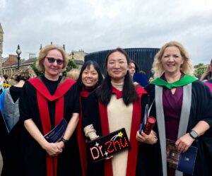 A group of people in academic robes, smiling at a graduation ceremony. One person holds a sign reading "Dr. Zhu." The mood is joyful and celebratory.